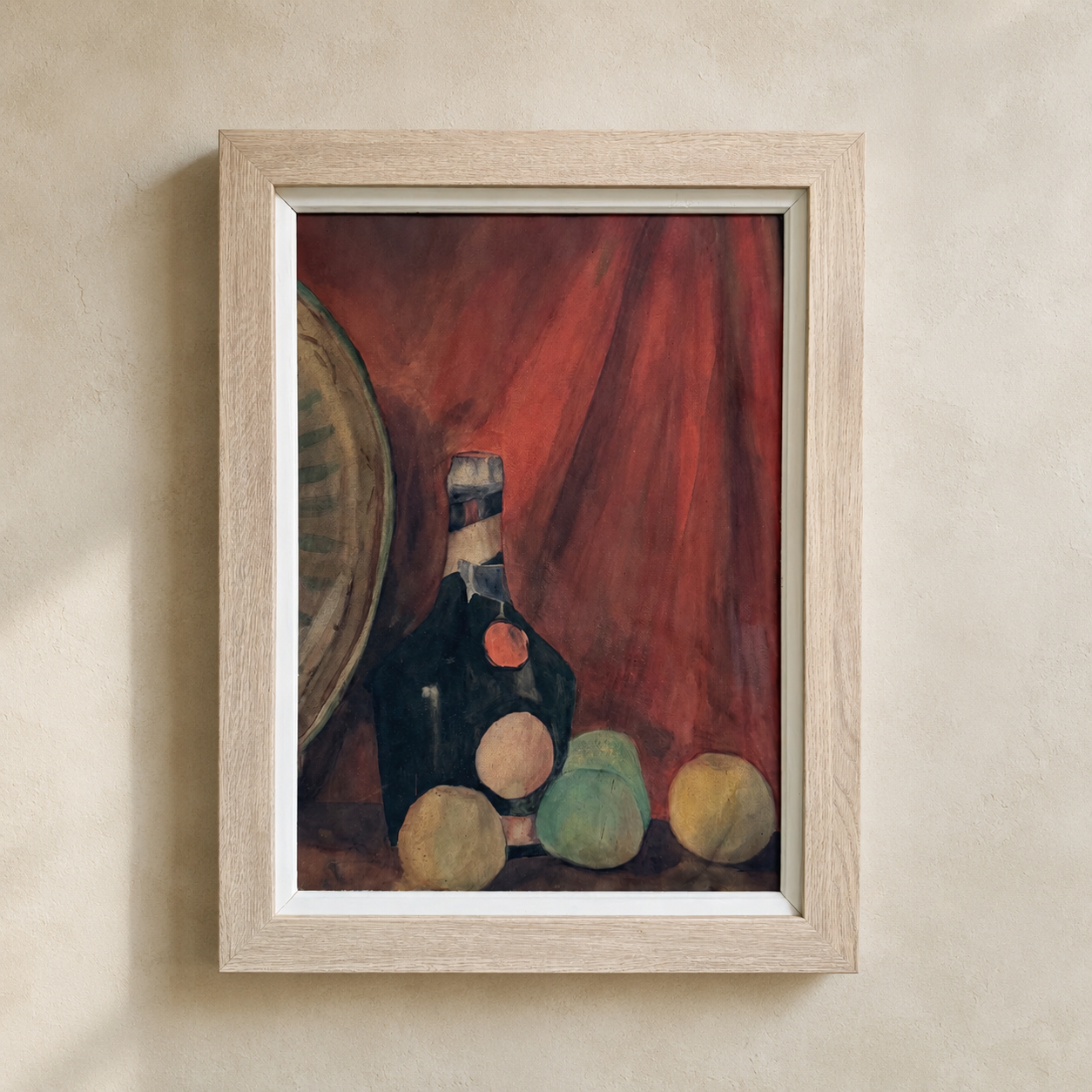 A framed still life painting featuring a dark bottle, green and yellow fruits, and a red curtain background, housed in a light natural wood frame and mounted on a textured beige wall.