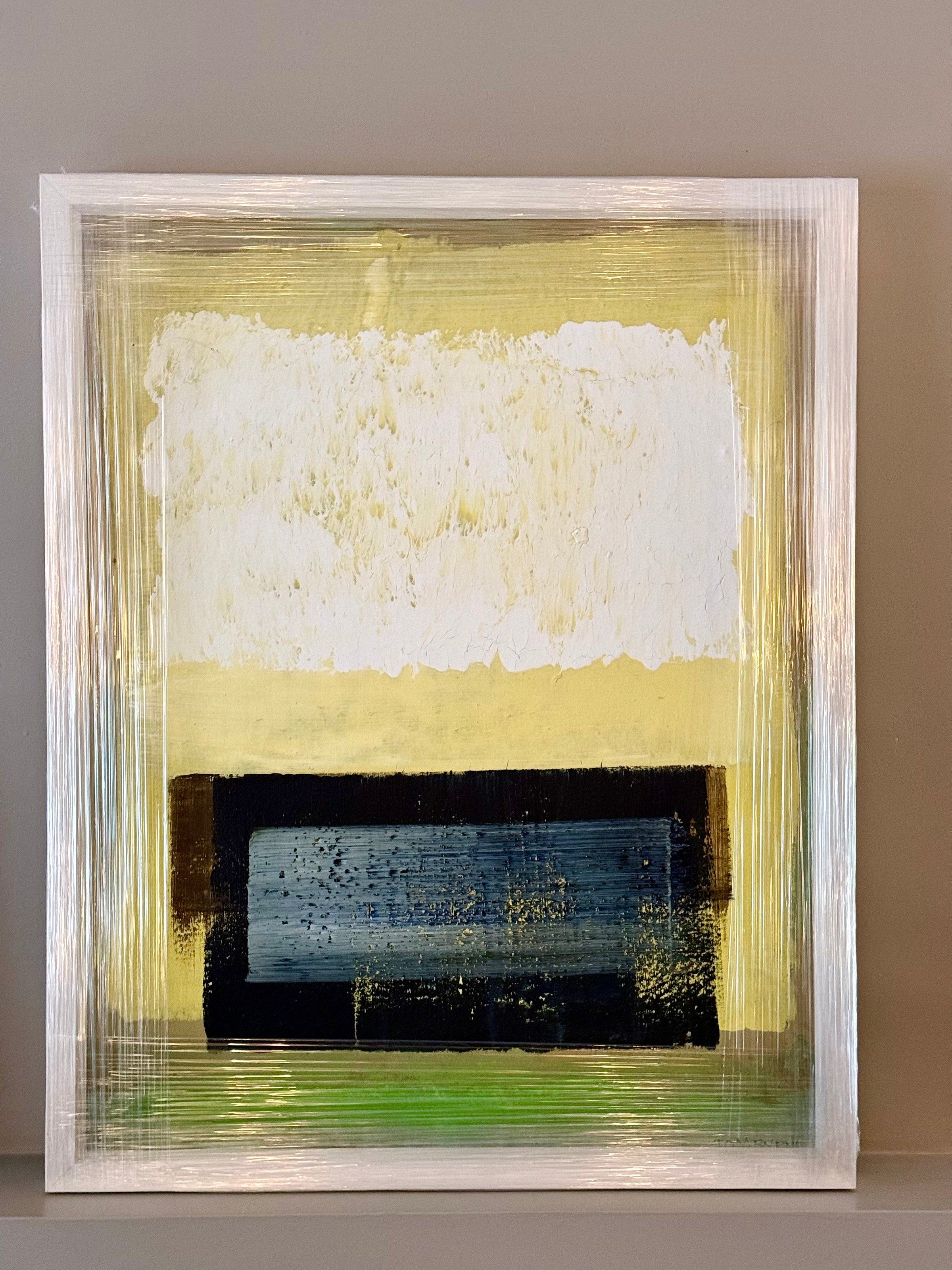 An abstract framed painting featuring a textured white block on a pale yellow background, with a dark navy and black rectangular section near the bottom and a thin green horizontal band. The artwork is housed in a wide, white textured frame and is displayed against a neutral gray wall.