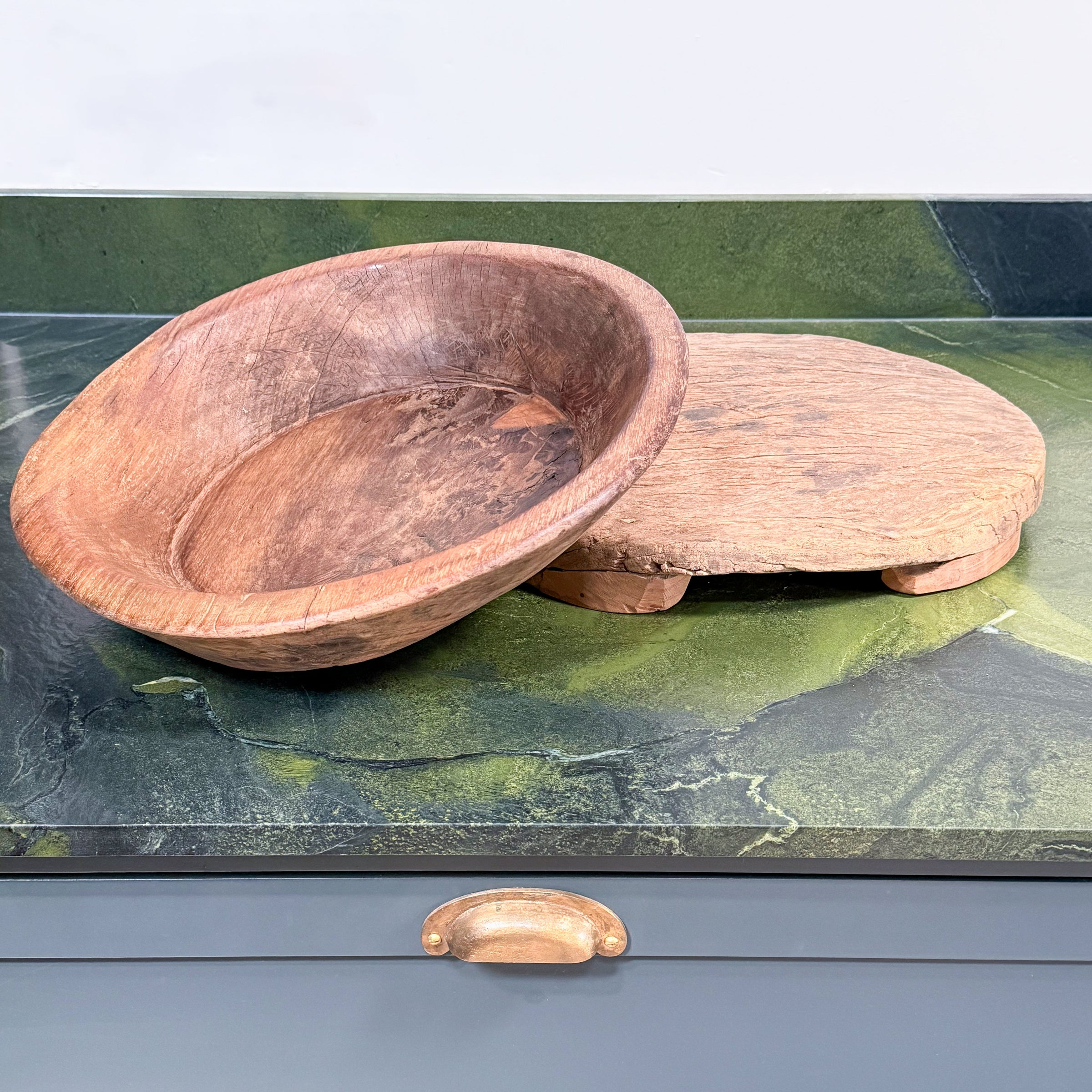 A rustic oval wooden bowl with a raised rim and a matching round wooden tray or lid, displayed on a green marble countertop above a blue-gray cabinet with a brass pull.