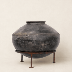 A large round dark ceramic vase with a weathered charcoal finish, displayed on a rusted metal stand against a plain beige background.