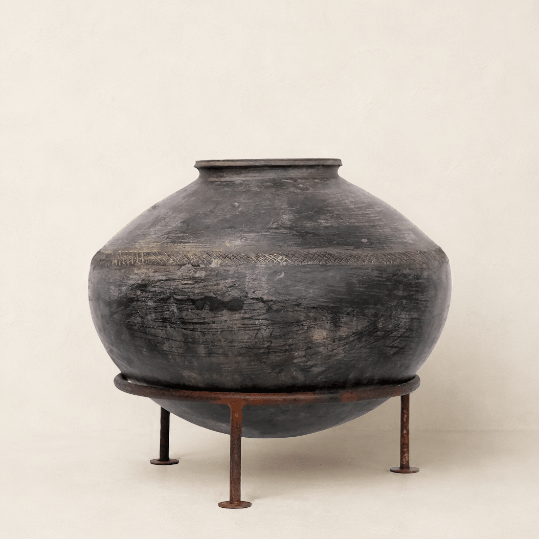 A large round dark ceramic vase with a weathered charcoal finish, displayed on a rusted metal stand against a plain beige background.