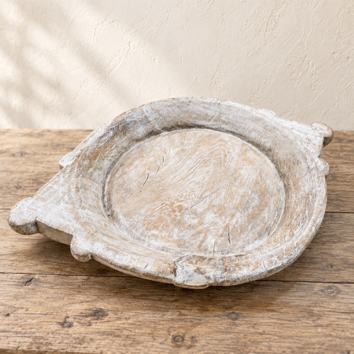 A rustic, round wooden tray with a distressed white and beige finish, featuring small side tabs, displayed on a weathered wooden table against a textured beige wall.