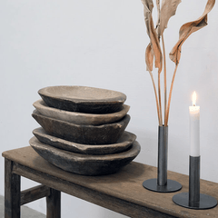 A stack of five rustic hand-carved wooden bowls in varying sizes sits on a weathered wooden table, styled next to a black metal candlestick holder with a lit white candle and dried palm leaves against a plain light gray wall.