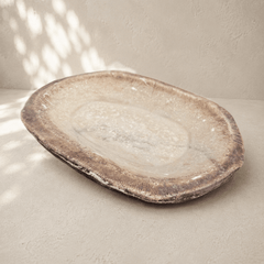 An organic-shaped stone serving platter with a textured, earthy brown and beige surface, photographed on a neutral concrete surface with soft dappled shadows.