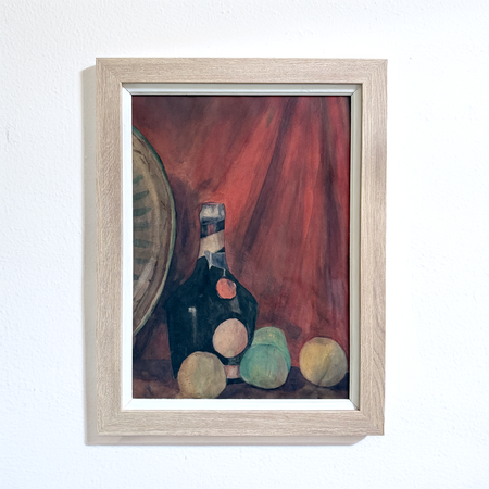A framed still life painting featuring a dark bottle, round fruits, and a red draped background, housed in a light natural wood frame and mounted on a white wall.