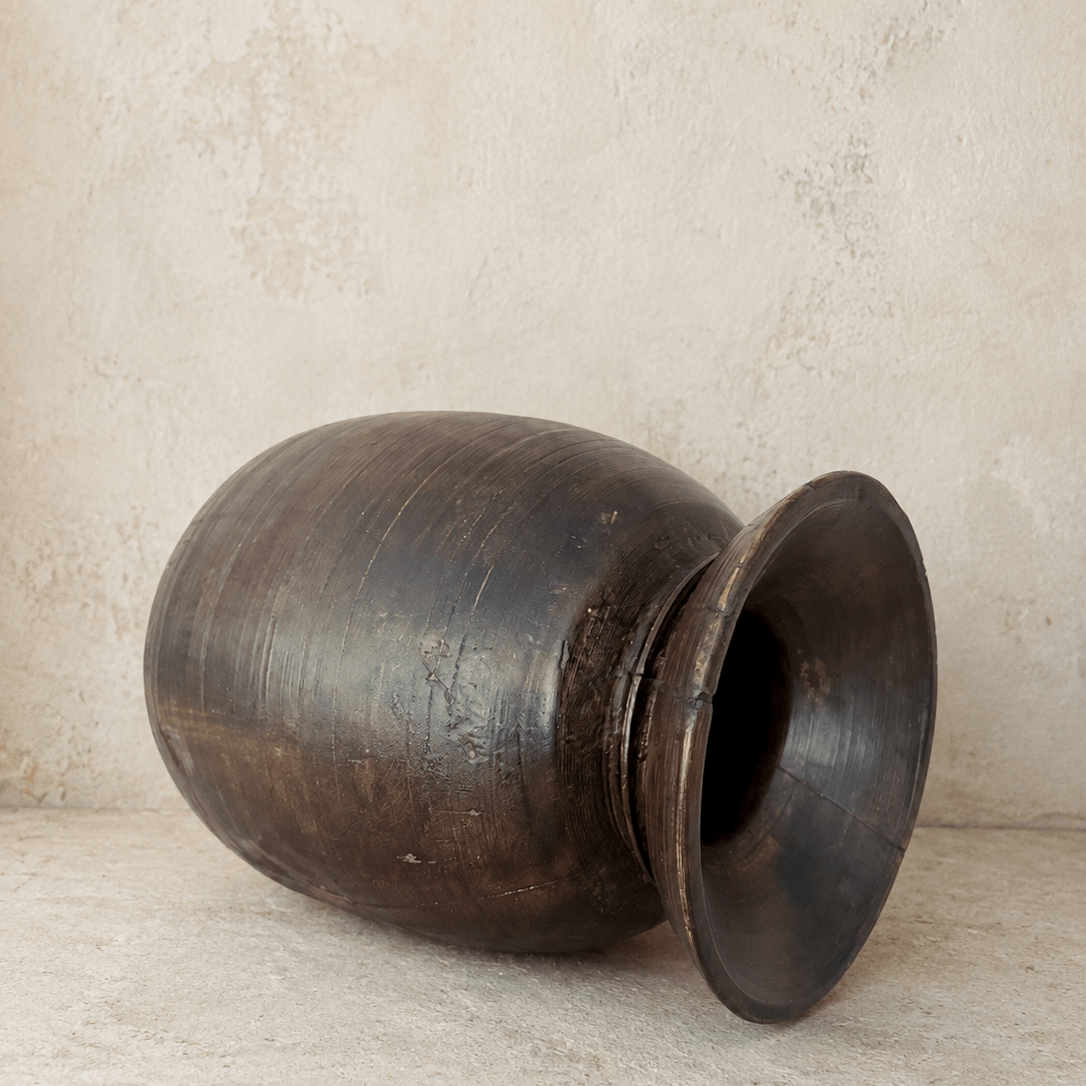 A large, dark brown wooden jar with a wide flared rim, lying on its side against a textured beige plaster wall and floor.