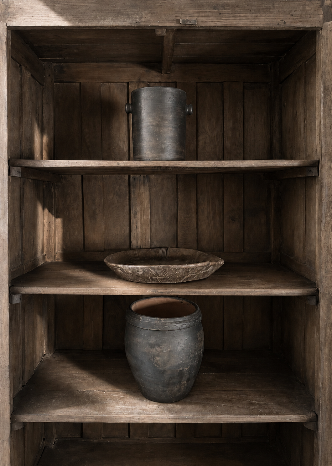 Three rustic dark storage canisters and a shallow wooden bowl displayed on three wooden shelves inside a matching wooden cabinet.