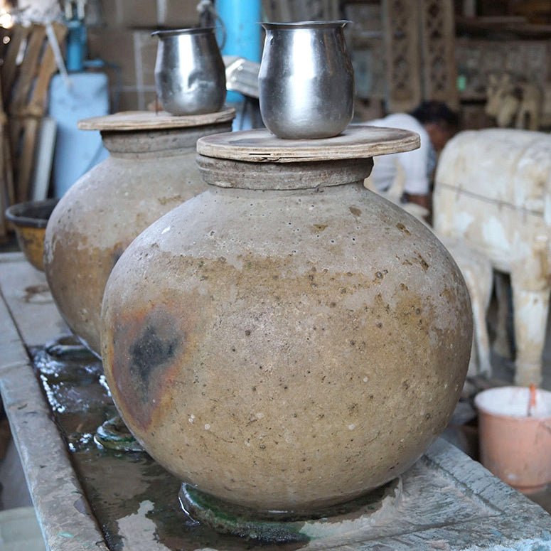 Three large round clay water pots with metal cups on top, arranged on a stone surface in a workshop setting.