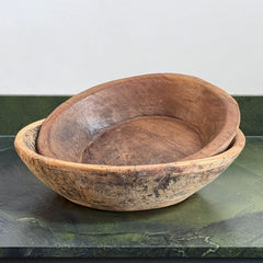 Two nested handcrafted wooden bowls with a rustic, weathered finish, displayed on a dark green marble surface against a light gray wall.