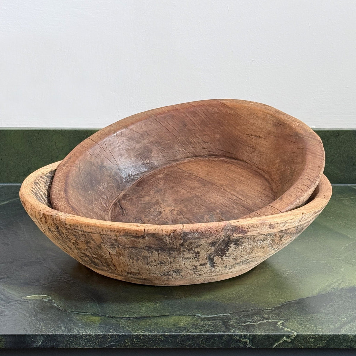 Two nested handcrafted wooden bowls with a rustic, weathered finish, displayed on a dark green marble surface against a light gray wall.