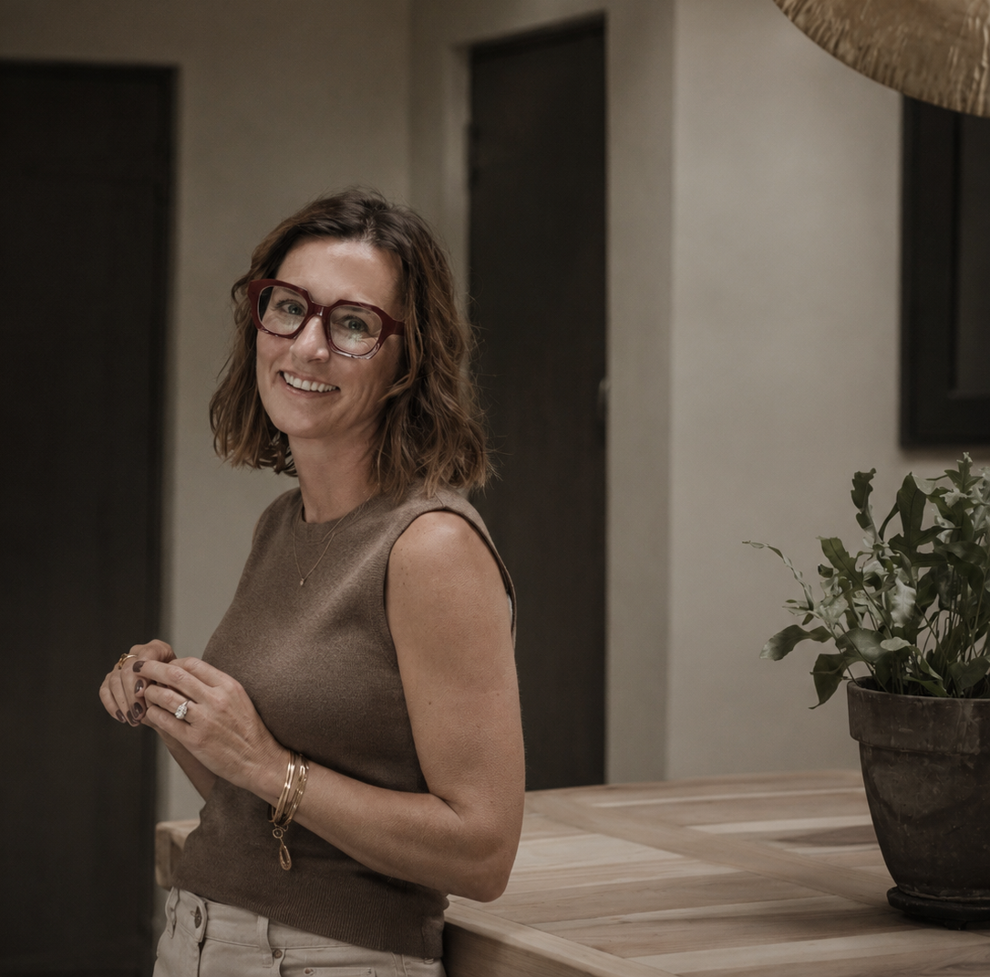 Woman wearing glasses and a sleeveless top, standing indoors with a plant in the background.