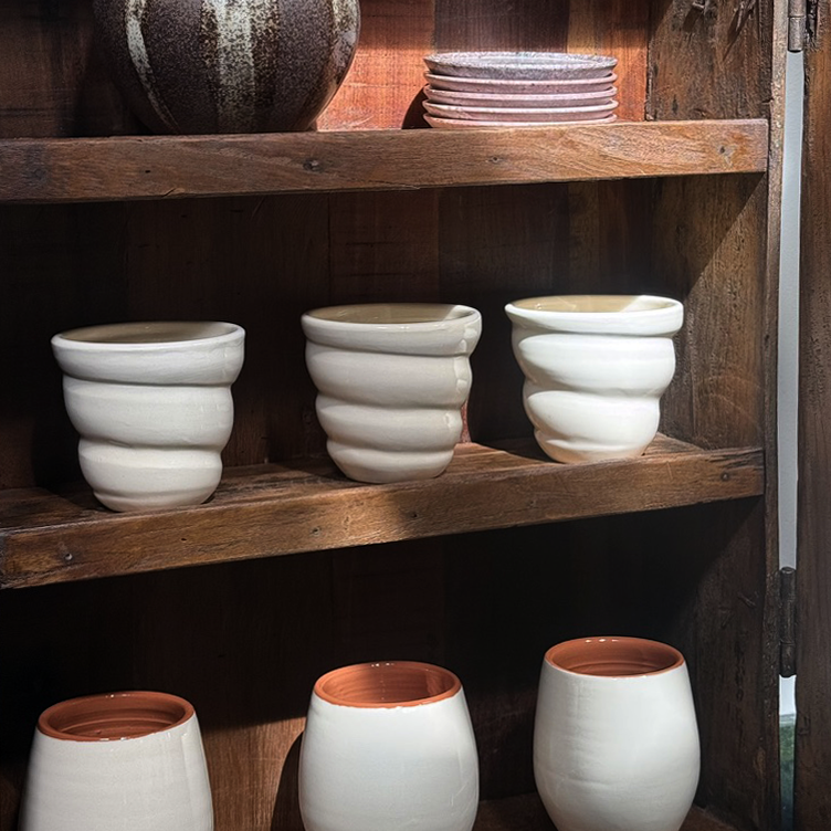 A set of handmade ceramic cups and small bowls with a matte off-white exterior and a warm terracotta interior, displayed on rustic wooden shelves.