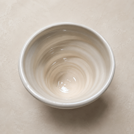 A small ceramic bowl with a glossy finish, featuring a swirling marbled pattern in cream and off-white tones, photographed from a top-down perspective on a textured beige surface.