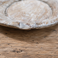 A close-up of a rustic, distressed ceramic plate or shallow dish with a heavily weathered, whitewashed finish showing wood grain underneath, resting on a matching rustic wooden surface.