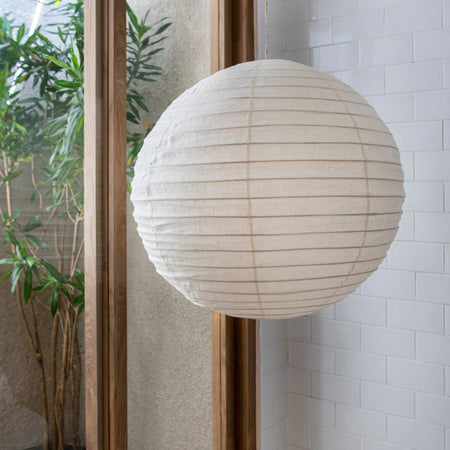 A large spherical off-white linen lantern hangs in front of a glass door frame, with a white tiled wall on the right and a potted plant visible outside on the left.