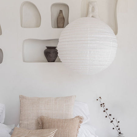 A large white linen lantern-style pendant light hangs in front of a white wall with recessed shelves holding small vases. Below, neutral beige and white pillows are arranged on a bed or sofa, with a small dried stem in a vase at the lower right.