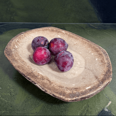 A rustic oval stone dish with a rough natural edge, holding four plums on a dark green textured surface.