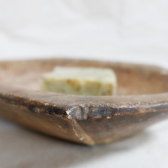 A rustic, hand-carved stone bowl with a thick, textured brown finish, holding a small, pale greenish-yellow rectangular object in the center, set against a soft, out-of-focus light background.