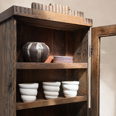 A rustic dark wood wall cabinet with an open door, displaying ceramic white bowls, cups, and a textured vase on its shelves against a neutral beige wall.