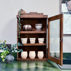A rustic wooden display cabinet with a glass-paneled door stands open on a green countertop, holding several cream-colored ceramic cups and bowls. A small potted eucalyptus plant sits to the left, and part of a framed artwork is visible on the wall behind.
