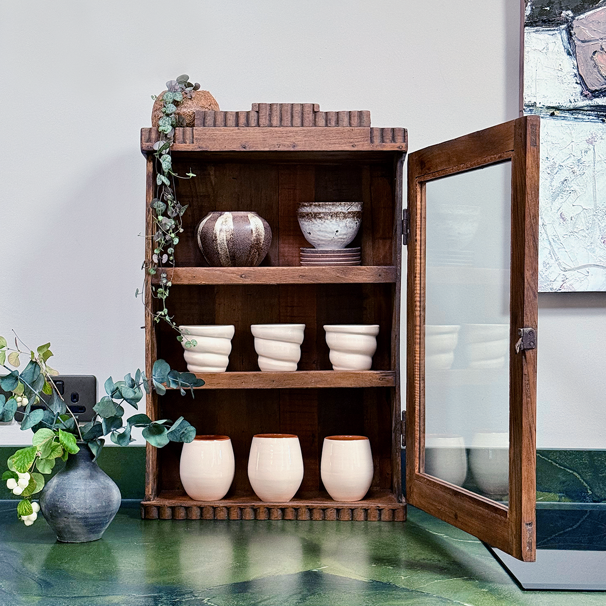 A rustic wooden display cabinet with a glass-paneled door stands open on a green countertop, holding several cream-colored ceramic cups and bowls. A small potted eucalyptus plant sits to the left, and part of a framed artwork is visible on the wall behind.