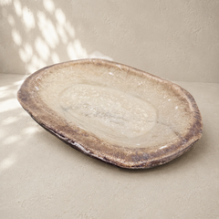 An organic-shaped stone serving platter with rustic, earthy brown, beige, and gray tones, set against a textured beige wall with soft dappled shadows.