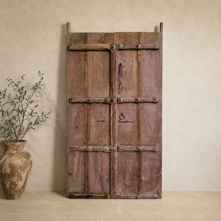 Vintage wooden door with metal handles against a beige wall with a plant on the left.