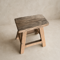 A rustic wooden stool with a weathered dark brown seat and natural wood legs, set against a neutral textured beige background.