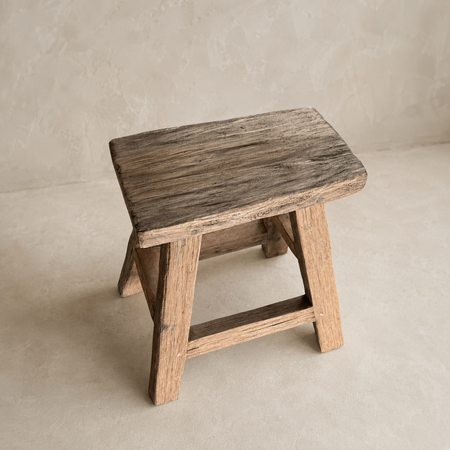 A rustic wooden stool with a weathered dark brown seat and natural wood legs, set against a neutral textured beige background.