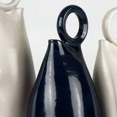 A glossy dark navy blue ceramic vase with a loop handle, shown in a close-up studio shot with other cream-colored vases partially visible on either side.