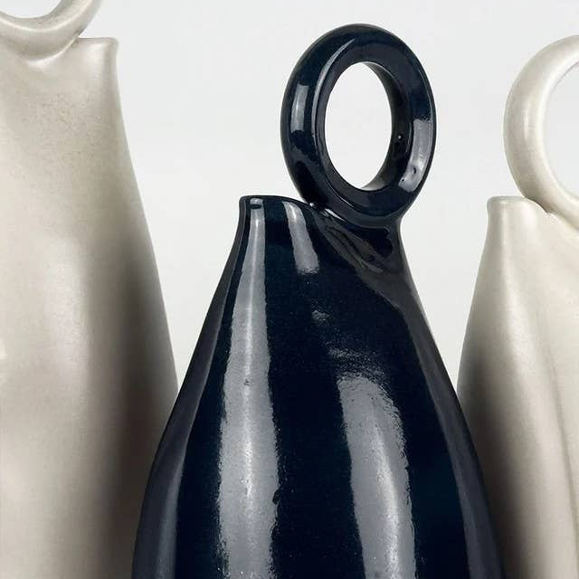 A glossy dark navy blue ceramic vase with a loop handle, shown in a close-up studio shot with other cream-colored vases partially visible on either side.