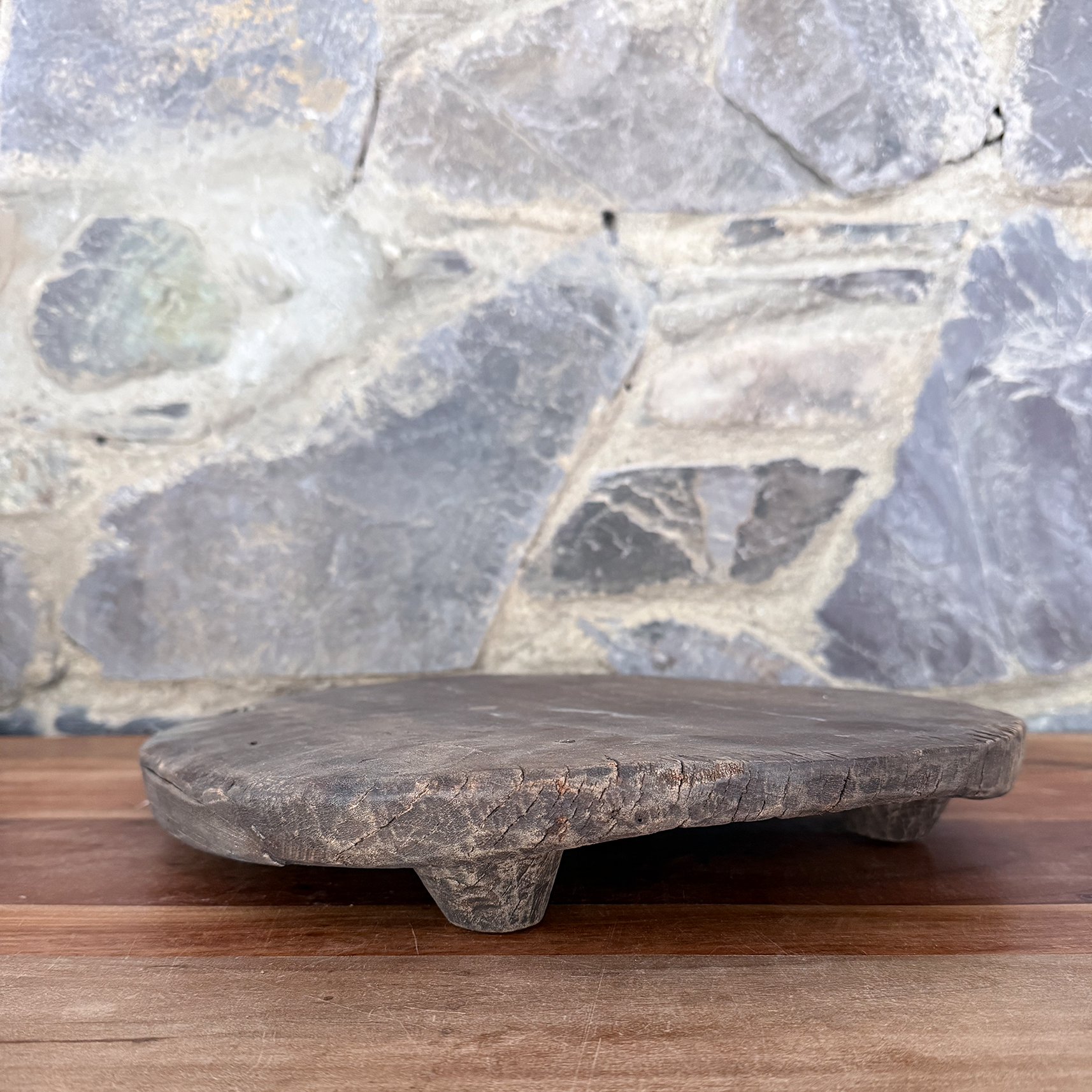 A rustic dark gray stone serving board with three short legs, resting on a wooden surface against a gray stone wall background.